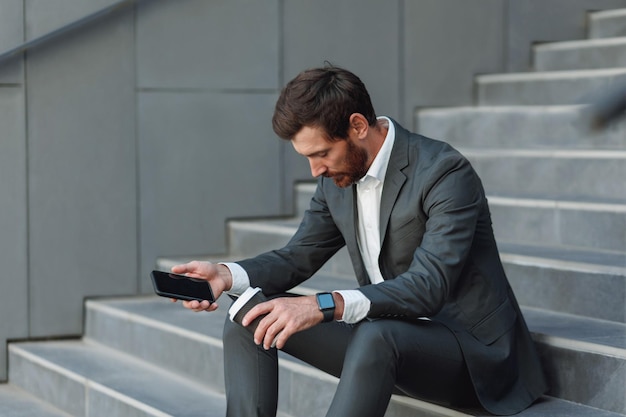 Homme d'affaires fatigué en costume avec téléphone et café pendant la pause assis dans les escaliers près du bureau