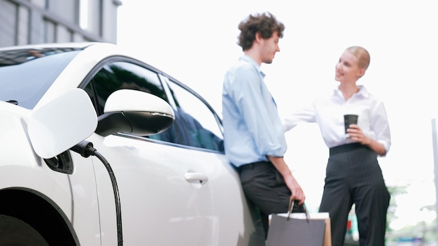 Un homme d'affaires et une femme d'affaires progressistes s'appuient sur une voiture électrique connectée à une station de charge avant de conduire dans le centre-ville une voiture rechargeable écologique alimentée par de l'énergie propre