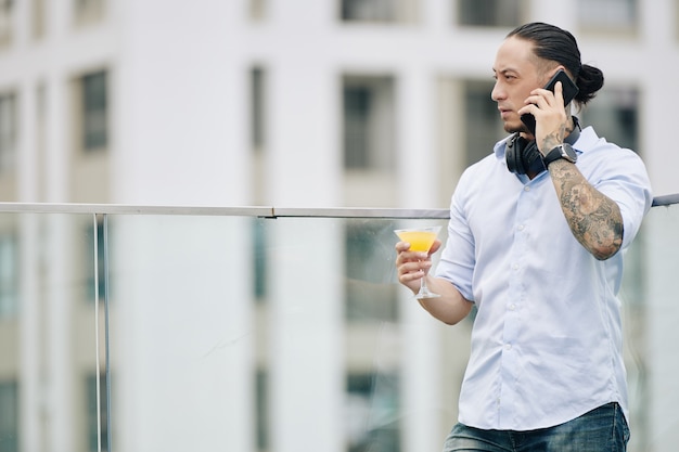 Homme d'affaires fronçant les sourcils parlant au téléphone en se tenant sur le toit et en savourant une savoureuse boisson à l'orange et en regardant le paysage urbain