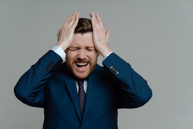 Homme d'affaires frustré en costume se sentant stressé et inquiet des problèmes financiers, un gars se touchant la tête avec les mains tout en recevant de mauvaises nouvelles en se tenant debout sur fond gris. Concept d'échec d'entreprise