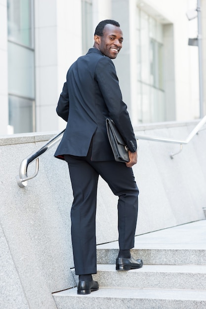 Homme d'affaires gai. Joyeux jeune homme africain en tenue de soirée se déplaçant par escalier et regardant par-dessus l'épaule