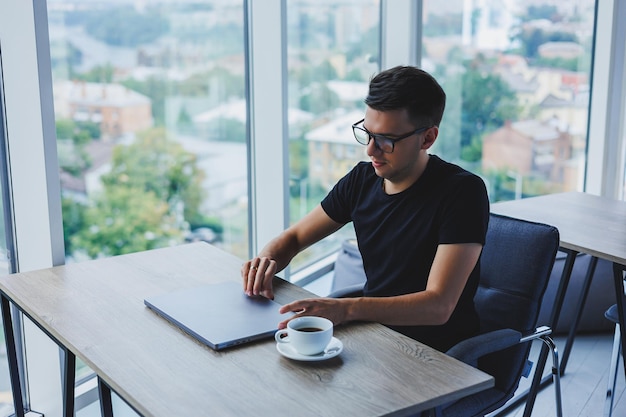 Un homme d'affaires gai à lunettes est assis à son bureau et travaille avec un ordinateur portable au bureau Un homme d'affaires est assis et travaille sur un ordinateur portable et boit du café Commencer la journée au bureau