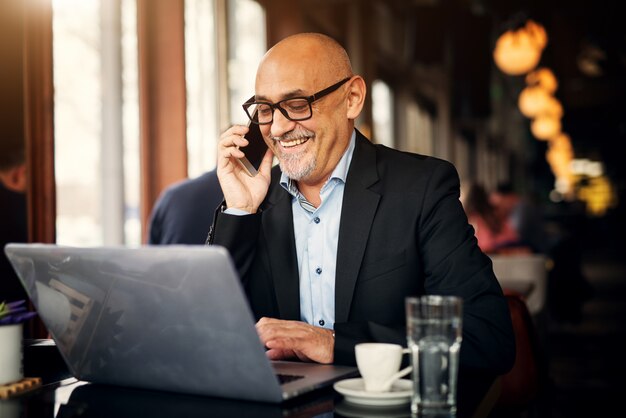 Homme d'affaires gai mature fait un appel téléphonique et utilise l'ordinateur portable tout en étant assis à la table d'un café et en riant