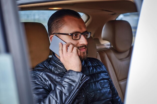 Homme d'affaires habillé à la mode assis sur un siège arrière dans la voiture de luxe, parlant par téléphone