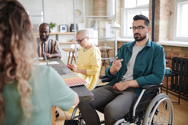 Homme d'affaires handicapé assis en fauteuil roulant et faisant des gestes, il travaille avec ses collègues lors d'une réunion d'affaires au bureau