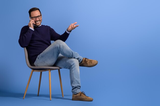 Photo un homme d'affaires heureux discutant d'idées sur son téléphone portable alors qu'il est assis sur une chaise sur un fond bleu