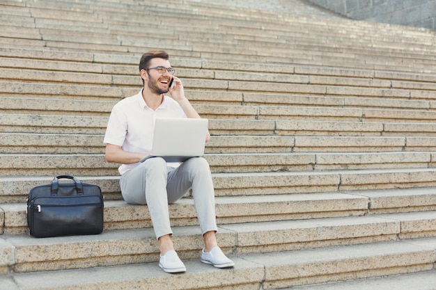 Un homme d'affaires heureux à lunettes a une conversation agréable au téléphone et travaille sur un ordinateur portable tout en étant assis sur des escaliers en pierre à côté de sa mallette. Technologie, communication et concept d'entreprise