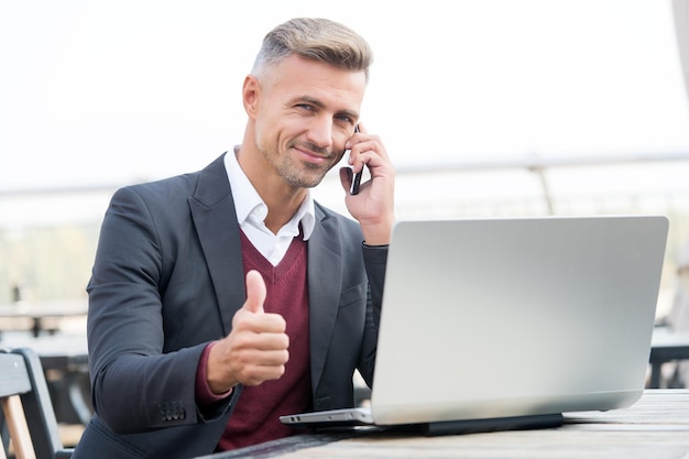 Un homme d'affaires heureux montre un geste de la main en parlant au client sur un téléphone portable et en utilisant un ordinateur pour l'approbation en ligne de l'entreprise