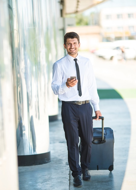 L'homme d'affaires heureux téléphone et marche avec une valise près du bâtiment