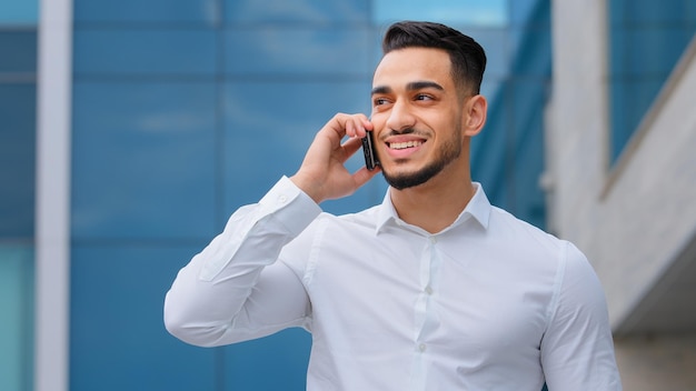 Un homme d'affaires hispanique indien barbu avec un téléphone portable dans la rue souriant un homme d'affaire arabe directeur répond à l'appel avec un smartphone à l'extérieur communication à distance un type joyeux parlant au téléphone portable à l'intérieur