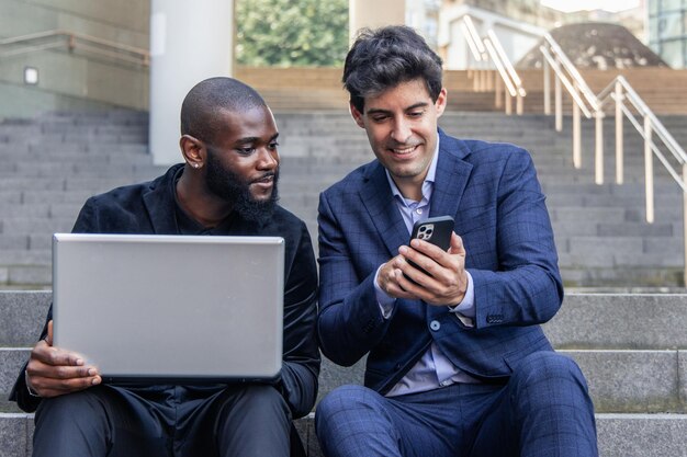 Un homme d'affaires hispanique montre les données de l'entreprise sur son smartphone à un collègue africain avec un ordinateur portable