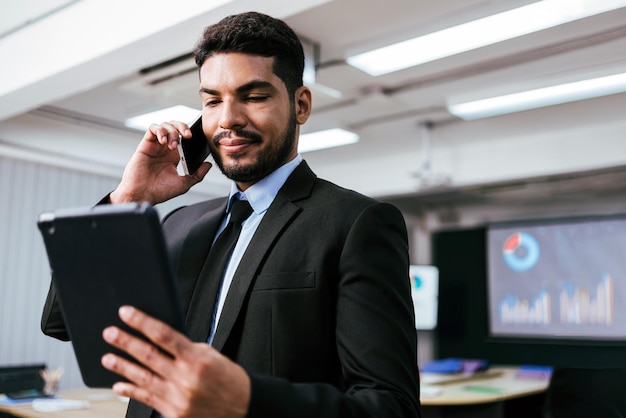 Un homme d'affaires intelligent et positif appelle et parle seul sur un smartphone tout en regardant une tablette numérique au bureau Un professionnel passe des appels professionnels et profite d'une conversation mobile d'entreprise à l'intérieur