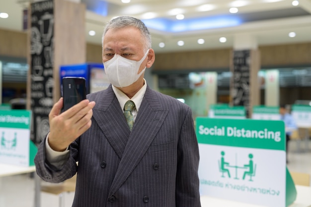 Homme d'affaires japonais mature avec masque à l'aide de téléphone et de la distance sociale à l'aire de restauration