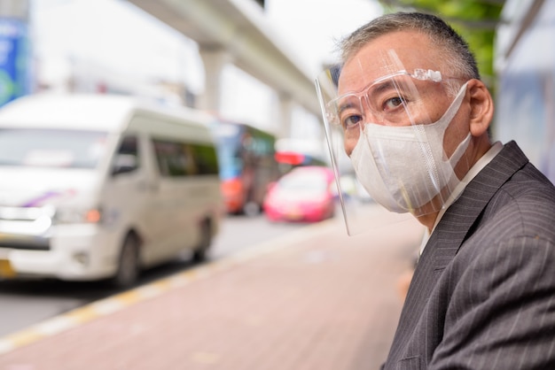 Homme d'affaires japonais mature avec masque et masque facial assis à l'arrêt de bus