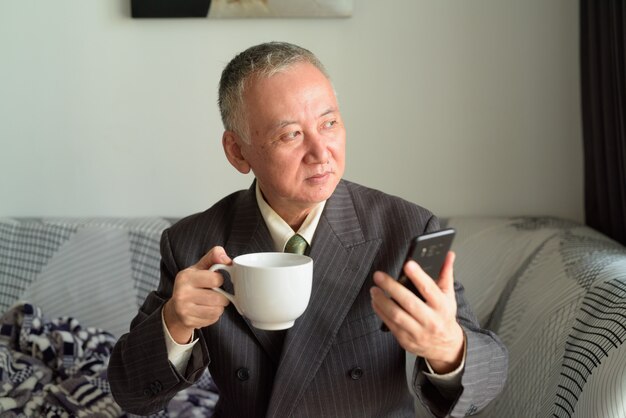 Homme d'affaires japonais mature pensant tout en tenant le téléphone et le café à la maison