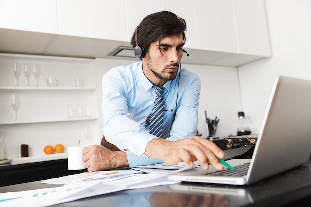 Homme d'affaires jeune concentré à la cuisine portant des écouteurs travaille avec des documents à l'aide d'un ordinateur portable.