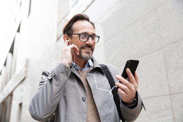 homme d'affaires joyeux à lunettes utilisant un téléphone portable et des écouteurs tout en se tenant près d'un immeuble de bureaux
