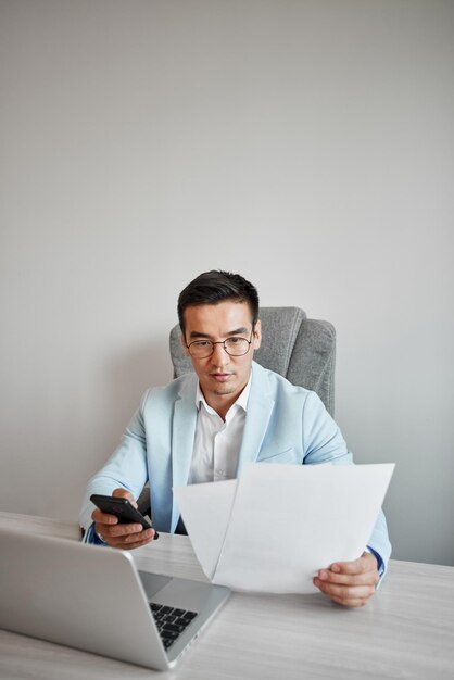 Un homme d'affaires kazakh asiatique en costume et lunettes avec un ordinateur portable au bureau parle par téléphone portable, résout des problèmes commerciaux, négocie un accord.