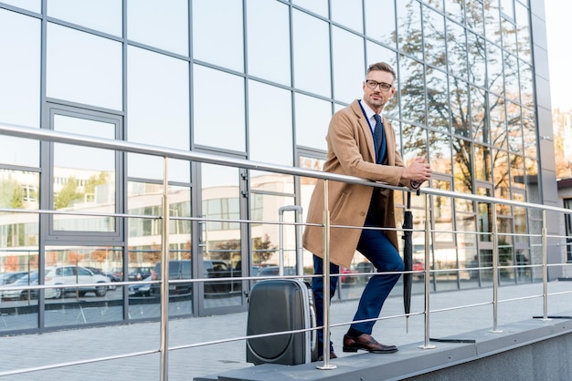 Homme d'affaires à lunettes et manteau beige debout avec parapluie près de valise