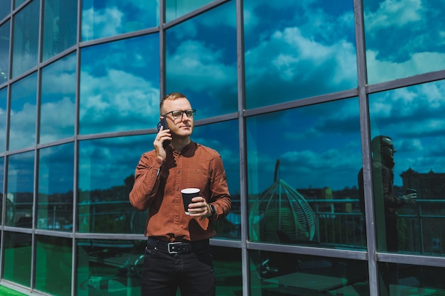 Homme d'affaires masculin à la mode attrayant dans une chemise et un pantalon élégants parlant au téléphone près de l'immeuble de bureaux et buvant du café bureau en verre moderne