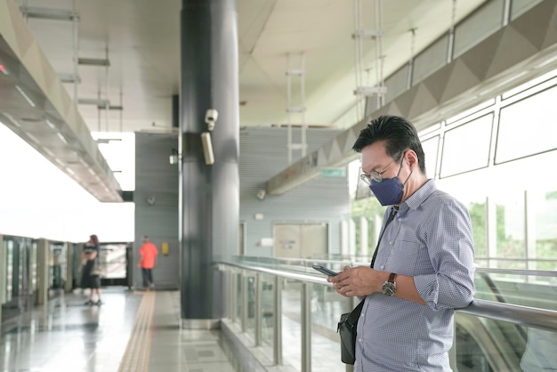 Homme d'affaires avec masque facial sur l'utilisation de son smartphone et attendant sur le quai du train