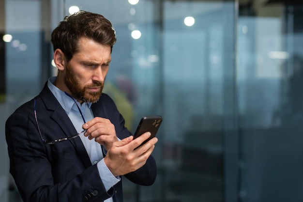 Homme d'affaires mature au bureau près de la fenêtre le soir contrarié par la lecture d'un message de notification en ligne