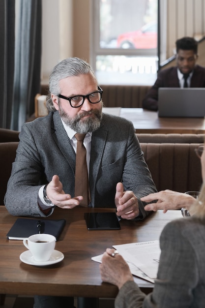 Homme d'affaires mature barbu assis à table avec son collègue et discutant du travail lors d'une réunion au restaurant