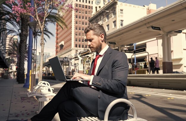 Homme d'affaires mature en costume assis sur un banc dans une ville en plein air travaillant en ligne sur un ordinateur portable indépendant