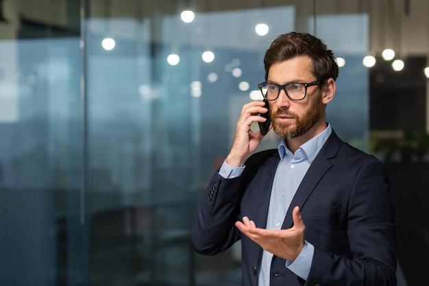 Homme d'affaires mature sérieux et prospère à l'intérieur du bureau près de la fenêtre parlant au téléphone homme d'affaires