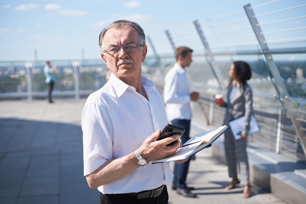 Homme d'affaires mature avec un smartphone et un ordinateur portable debout sur la terrasse d'un centre d'affaires