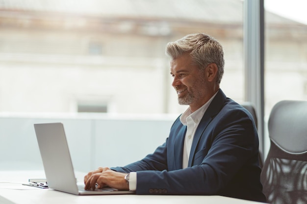 Homme d'affaires mature souriant travaillant sur un ordinateur portable assis dans un coworking moderne