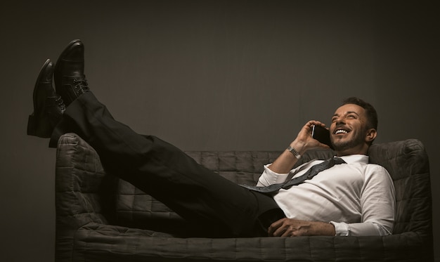 Homme d'affaires avec mobile se trouve jeter les jambes sur le canapé. Homme souriant allongé sur le canapé, les jambes croisées, parler au smartphone sur le dos gris.
