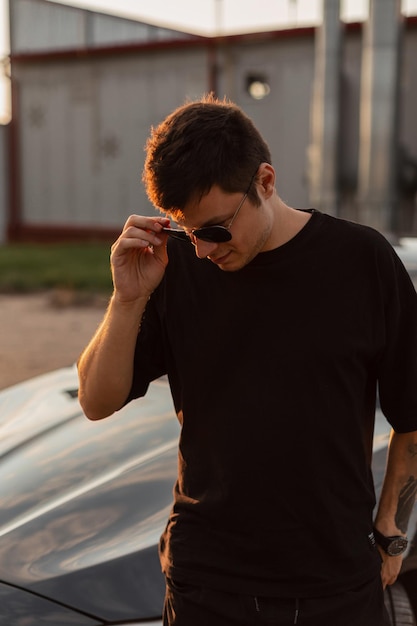 Homme d'affaires de mode en vêtements noirs marche près d'une voiture et met des lunettes de soleil élégantes au coucher du soleil