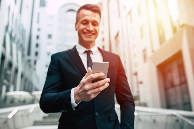 Homme d'affaires moderne, beau et confiant dans une tenue de soirée utilise un téléphone intelligent à l'extérieur