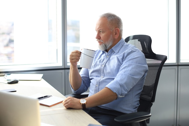 Homme d'affaires mûr concentré plongé dans ses pensées alors qu'il était assis au bureau avec une tasse de café à la main dans un bureau moderne.