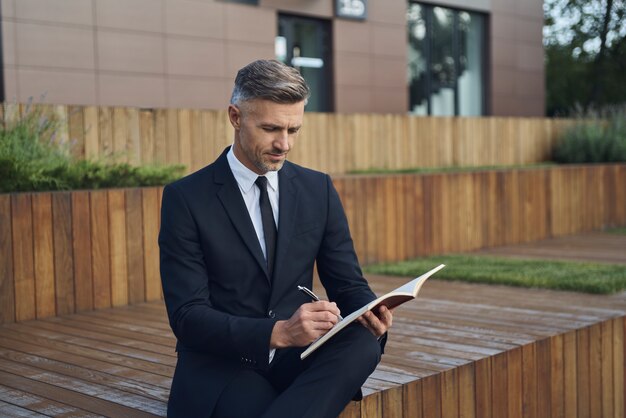 Homme d'affaires mûr et confiant prenant des notes tout en étant assis à l'extérieur près d'un immeuble de bureaux