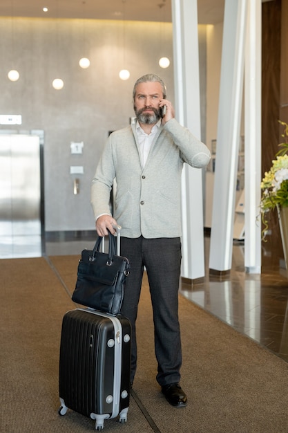 Homme d'affaires mûr avec une valise parlant au téléphone portable dans le salon de l'hôtel