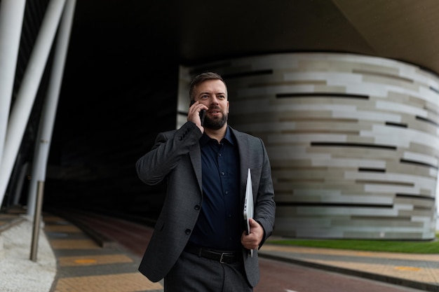 Homme d'affaires avec un ordinateur portable sous le bras parlant au téléphone tout en marchant le long du bâtiment