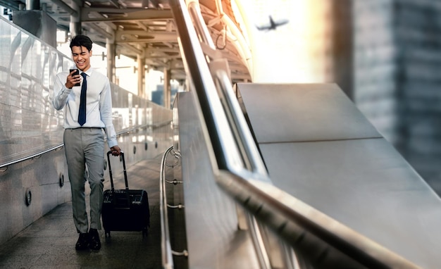 Homme d'affaires passager souriant utilisant un téléphone portable tout en marchant avec une valise à l'aéroport pour un voyage d'affaires sur toute la longueur