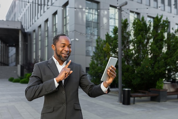 Homme d'affaires de patron afro-américain mature à l'extérieur de l'immeuble de bureaux avec casque et tablette parlant