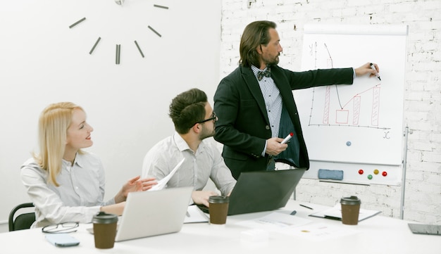 Homme d'affaires patron se tient près d'un tableau blanc présentant à l'équipe une nouvelle idée d'entreprise. Collègues écoutant le haut-parleur assis au bureau.