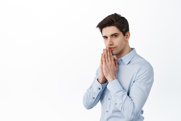 Homme d'affaires PDG, homme d'affaires priant, se tenant la main en prière, regardant devant avec anticipation et visage sérieux, mendiant, debout sur un mur blanc