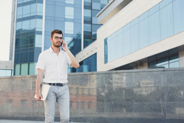 Un homme d'affaires pensif parle sur un téléphone portable et tient une tablette numérique dans sa main tout en se tenant devant un immeuble de bureaux moderne, copiez l'espace. Concept d'entreprise, de technologie et de communication