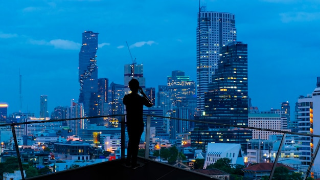 Homme d'affaires permanent à l'aide d'un téléphone intelligent sur le balcon ouvert sur le toit en regardant la vue nocturne de la ville. Affaires ambitieuse et vision.