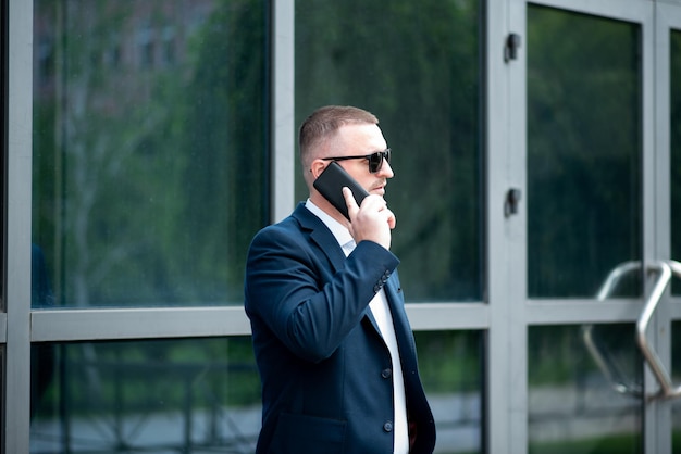 Un homme d'affaires portant des lunettes de soleil foncées se tient à l'extérieur du bâtiment de l'entreprise et parle au téléphone