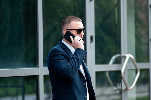 Un homme d'affaires portant des lunettes de soleil foncées se tient à l'extérieur du bâtiment de l'entreprise et parle au téléphone