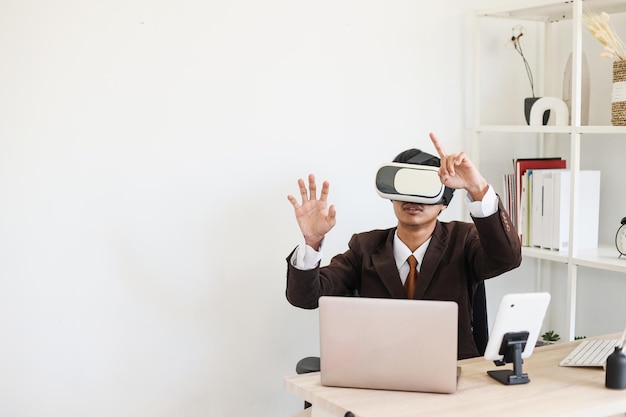 un homme d'affaires portant des lunettes VR est assis devant un ordinateur portable au bureau, gesticulant les mains.