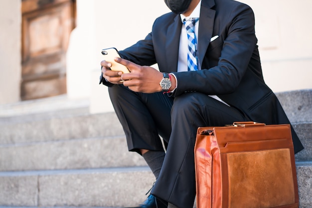 Homme d'affaires portant un masque facial et utilisant son téléphone portable alors qu'il était assis dans les escaliers à l'extérieur. Nouveau style de vie normal. Concept d'entreprise.