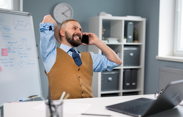 Homme d'affaires positif en vêtements élégants assis au bureau avec un ordinateur portable et parlant sur un homme barbu caucasien utilisant des gadgets modernes pour travailler au bureau