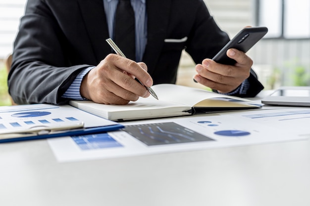 L'homme d'affaires prend des notes depuis son téléphone portable dans un cahier, il est propriétaire de l'entreprise, il vérifie les documents financiers de l'entreprise dans son bureau. Concept de gestion financière.